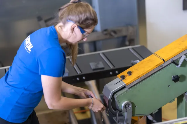 Mitarbeiterin mit Schutzbrille entgratet ein Werkstück am Bandschleifer, Funkenflug an der Schleifscheibe